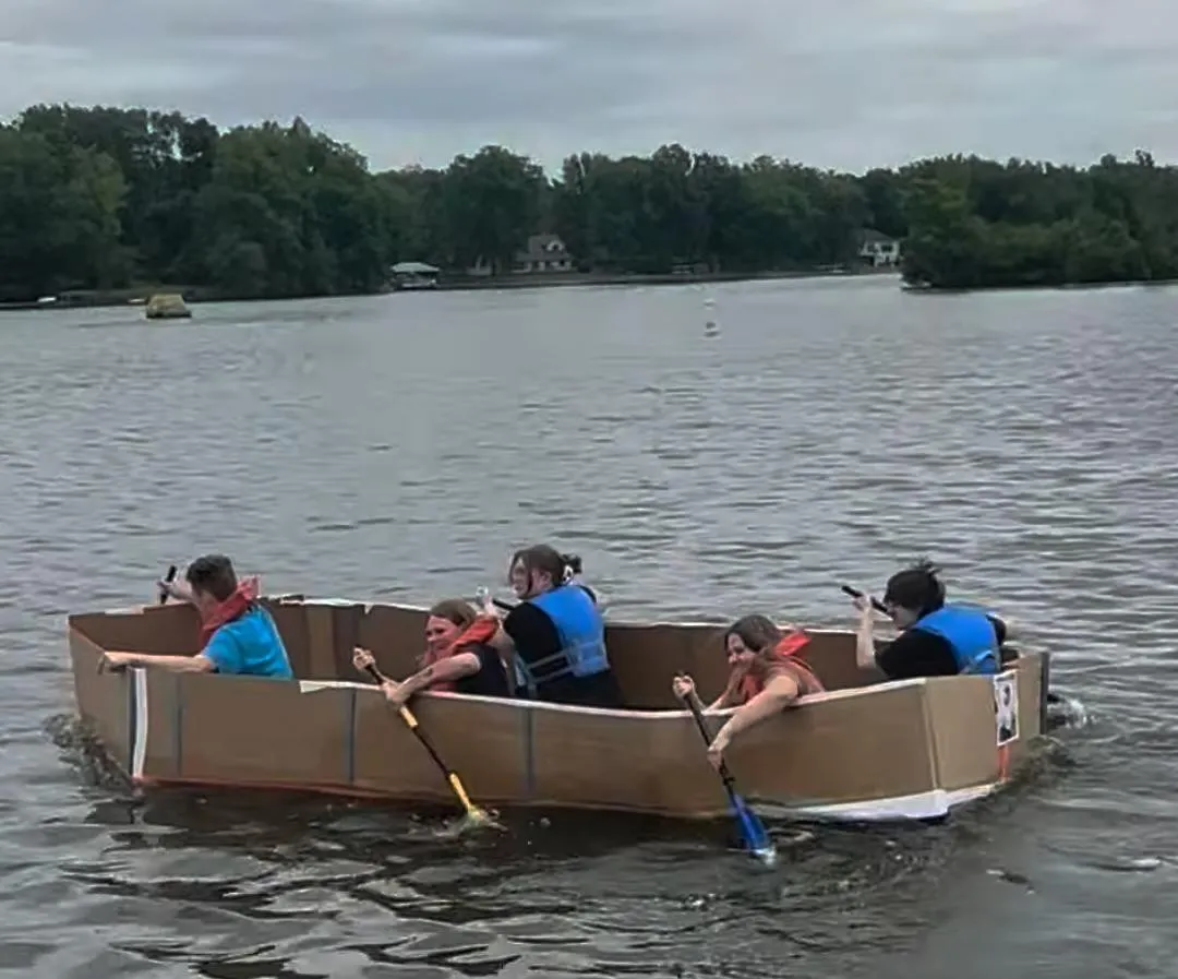 Five crew members paddling the 20-foot cardboard boat across a lake, all wearing life jackets