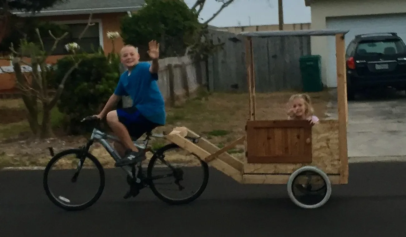The bike-drawn chariot on a neighborhood street: a wooden body on salvaged bicycle wheels under a tarp roof, towed behind a bicycle with a passenger riding inside